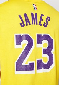 Gul NBA-tröja med "JAMES" i lila bokstäver ovanför en stor "23" i lila med vit kontur, tillverkad av mjukt bomullstyg.