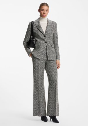 Young woman wearing a gray herringbone suit with wide-leg pants, white turtleneck, black handbag, and black high-heeled shoes standing against white background.