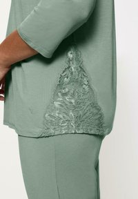 Top verde con una tela suave y mangas de 3/4, con un detalle de encaje en el lado en un patrón floral, añadiendo textura y diseño.