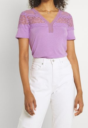 Kobieta ubrana w fioletowy krótki top z koronkowym detalem na wysokości klatki piersiowej, zestawiony z białymi spodniami z wysokim stanem.