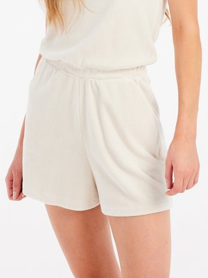 Vrouw draagt lichtbeige badstofshorts met een elastische tailleband en zakken, staand tegen een witte achtergrond.