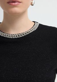 Haut en tricot noir avec un col décoratif en perles argentées et nacrées. Le tissu a un léger brillant, mettant en valeur le détail du décolleté.