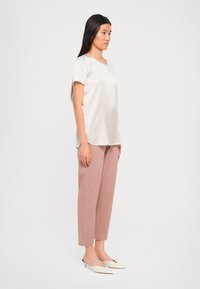 Una donna con lunghi capelli neri è in piedi di profilo, indossando una blusa corta in satin crema, pantaloni rosa polveroso alla caviglia e mule crema con tacco.