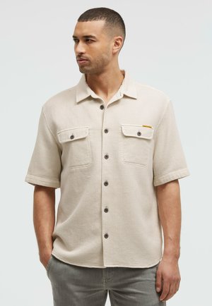 Mann mit kurzen Haaren, der ein beiges kurzärmeliges Hemd mit zwei Brusttaschen und graue Hosen trägt, steht mit den Händen in den Taschen.