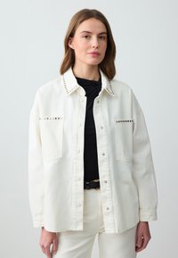 Veste en denim blanc avec un col clouté, comportant deux poches avant et une coupe décontractée. Portée sur un haut noir et associée à un pantalon blanc.
