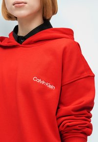 Άτομο που φοράει κόκκινο φούτερ Calvin Klein με κουκούλα πάνω από μαύρο πουκάμισο με γιακά, εμφανίζοντας τον άνω κορμό και το τσαλακωμένο μανίκι στο δεξί χέρι.