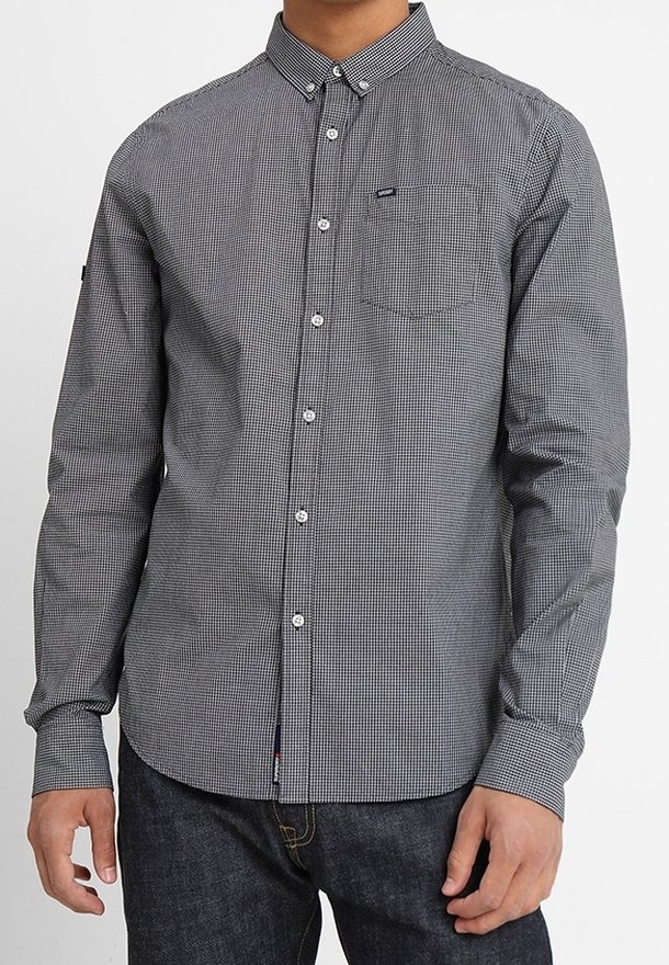 Homme portant une chemise à carreaux noire et blanche à manches longues, boutonnée avec une poche poitrine, et un jean foncé, debout devant un fond blanc.