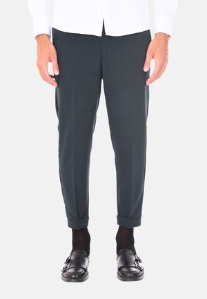 Pantaloni sartoriali verde scuro con una texture liscia, presentano un orlo arrotolato e lievi pieghe frontali, abbinati a mocassini neri e calzini scuri.