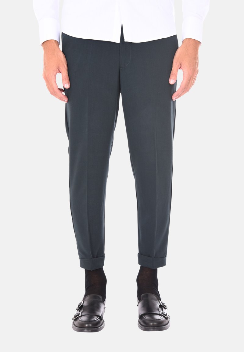Pantaloni sartoriali verde scuro con una texture liscia, presentano un orlo arrotolato e lievi pieghe frontali, abbinati a mocassini neri e calzini scuri.