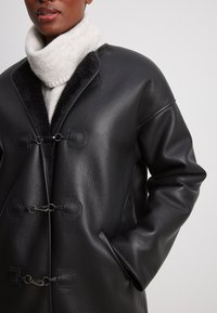 Juodas odinis paltas su tekstūruotu paviršiumi, apdailintas sidabro spalvos detalėmis, turintis dvi priekinės kišenes ir aukštą apykaklę, prieš neutralų foną.