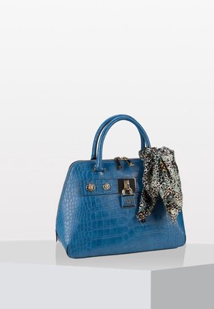 Bolso azul con estampado de cocodrilo con cierre y botones dorados, que lleva un pañuelo con estampado de leopardo atado a una de las asas, colocado sobre una superficie blanca.