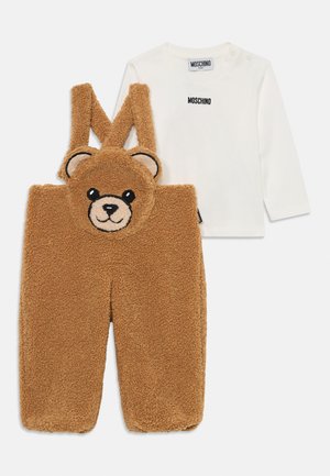 Salopette en polaire marron avec un visage d'ours en peluche, des yeux noirs et des oreilles. Chemise à manches longues blanche avec le logo "Moschino" en noir.