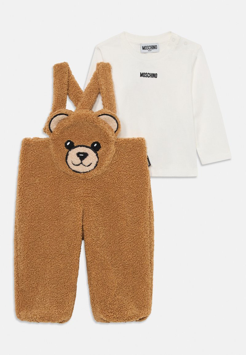 Bruine fleece overall met een teddybeer gezicht, zwarte ogen en oren. Witte shirt met lange mouwen met "Moschino" logo in het zwart.