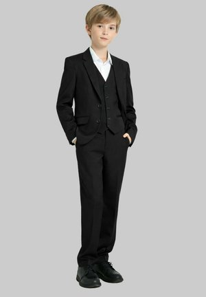 Jeune personne debout, les mains dans les poches, portant un costume trois pièces noir, une chemise blanche et des chaussures habillées noires sur un fond gris.