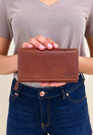 Brown leather wallet with stitched edges, featuring embossed logo. Held between hands, with a plain gray shirt and dark jeans partially visible.