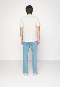 Man som står vänd mot en vit vägg, iförd en enfärgad vit t-shirt, ljusblå jeans och vita sneakers, med tatueringar på sina armar.