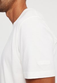 White cotton t-shirt featuring a crew neck and short sleeves, with a subtle embossed logo on the left sleeve. Soft texture.