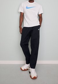 Hombre vistiendo una camiseta blanca de Nike con el swoosh azul, pantalones cargo negros de Nike y zapatillas blancas con suelas marrones, de pie contra una pared pálida.