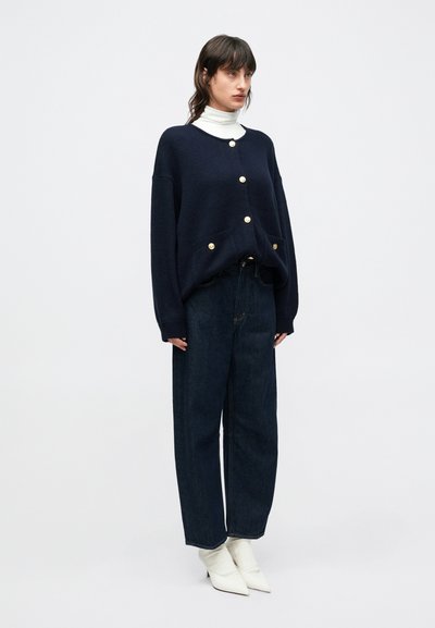 Tengerészkék kötött kardigán arany gombokkal, túlméretezett szabásban, magasderekú sötét farmerral és fehér, hegyes orr�ú csizmával párosítva. Sima textúra.