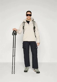 Light beige waterproof jacket with hood, black cargo pants, gray hiking shoes, and sunglasses. Holding black trekking poles.
