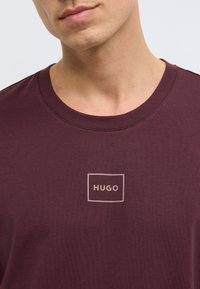 Μπορντό μπλουζάκι με στρογγυλή λαιμόκοψη που διαθέτει ένα διακριτικό λογότυπο μέσα σε ορθογώνιο πλαίσιο, κατασκευασμένο από απαλό ύφασμα με λεία υφή.