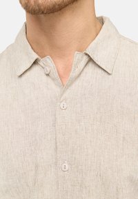 Camisa de lino beige claro con cuello, dos botones en la parte delantera y una tela texturizada con un sutil patrón de tejido.