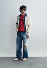 Uomo con occhiali da sole neri, cardigan bianco a maglia sopra una giacca di jeans, t-shirt rossa con stampa grafica, jeans larghi con toppe e sneakers bianche in piedi.