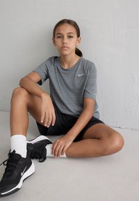 Grå atletisk t-shirt med striber og Nike-logo, sorte shorts, sorte sko med hvide accenter og hvide sokker. Sidder på en lys grå gulv.