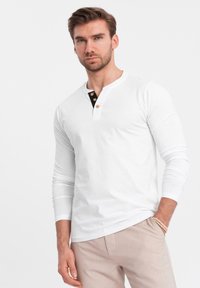Mann mit Bart, der ein weißes Langarm-Henley-Shirt mit Knöpfen und beigen Hosen trägt, steht mit einer Hand in der Tasche vor einem weißen Hintergrund.