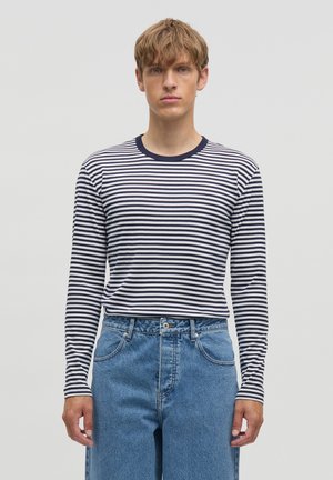 Giovane uomo con una camicia a maniche lunghe a righe blu navy e bianco infilata in jeans blu a vita alta, in piedi davanti a un semplice sfondo chiaro.