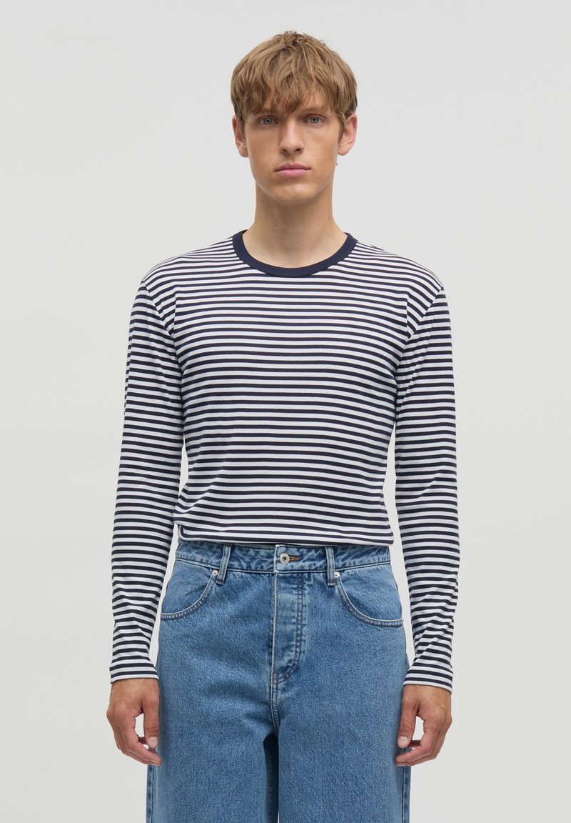 Mladý muž nosící námořnicky modré a bílé pruhované tričko s dlouhým rukávem zastrčené do modrých džínů s vysokým pasem, stojící před jednoduchým světlým pozadím.