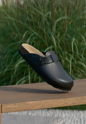 Zoccolo in pelle nera con punta arrotondata, cinturino regolabile con fibbia, soletta in sughero e suola in gomma testurizzata, esposto su una superficie di legno.