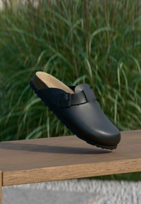 Zoccolo in pelle nera con punta arrotondata, cinturino regolabile con fibbia, soletta in sughero e suola in gomma testurizzata, esposto su una superficie di legno.