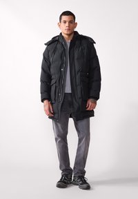 Parka acolchada negra con cierre de cremallera en frente y cierres a presión, con capucha desmontable y bolsillos laterales. Combinada con jeans grises y zapatillas.