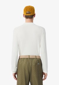 Maglietta bianca a maniche lunghe abbinata a pantaloni verdi oliva con motivo, dotati di una fascia elastica in vita e una cintura beige, completati da un berretto senape.