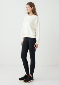Pullover crema con cuciture dorate, colletto rotondo e maniche lunghe, abbinato a leggings neri e sneakers nere, mostrato su uno sfondo semplice.