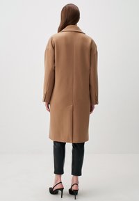 Lungo cappotto di lana beige con un ampio collo, spalle scese e dettagli con bottoni. Indossato sopra pantaloni neri aderenti e eleganti scarpe con tacco alto.