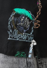 Camisa negra texturizada con un gráfico que presenta una calavera sosteniendo una guadaña, palmeras y detalles de cadena coloridos. Costuras no estándar visibles.
