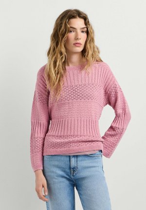 Jonge vrouw met golvend haar die een gestructureerde roze gebreide trui en lichtblauwe spijkerbroek draagt, staande tegen een effen lichte achtergrond.