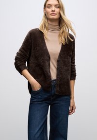 Brun, fluffig cardigan med knäppning, vida ärmar och en avslappnad passform, som matchas med en beige polotröja och blå jeans.