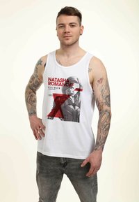 Hvid tanktop med grafisk print af Natasha Romanoff, med røde accenter og tekst. Modellen har tatoveringer og bærer slidte grå jeans.
