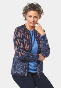 Cardigan marine avec des motifs floraux et géométriques dans des nuances de bleu, orange et rose. Il présente un col rond et une fermeture éclair à l'avant.