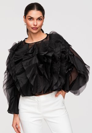 Kobieta o ciemnych włosach nosi przezroczystą czarną bluzkę z falbankami i białe spodnie, pozując z ręką na biodrze na neutralnym tle.