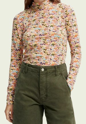 Mujer con una blusa floral de manga larga y cuello alto, metida dentro de unos pantalones de tiro alto de color verde oliva, con la mano izquierda en el bolsillo.