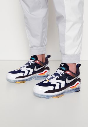 Une personne porte un pantalon de jogging blanc et des baskets Nike Air Max noires, blanches et orange avec des semelles transparentes, debout sur un sol blanc.