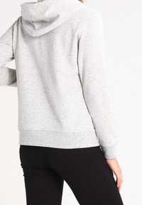Sudadera gris con una ligera textura, que presenta una capucha y puños acanalados. El diseño es casual con un ajuste de longitud media. Combinada con pantalones negros.