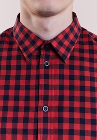 Camisa a cuadros rojos y marinos con cuello clásico, cierre de botones en la parte delantera y tejido texturizado. Presenta botones negros y un patrón de espiga.