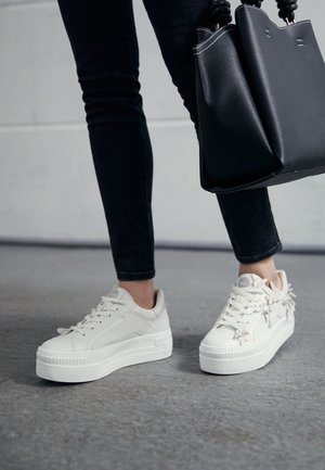 Weiße Plateausneaker mit strukturierten Gummisohlen, verziert mit Schnürsenkel-Details. Kombiniert mit dunklen Skinny-Jeans und einer schwarzen Handtasche.