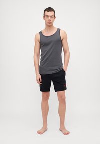 Un maieu gri asortat cu pantaloni scurți negri care au un patch cu logo roșu. Materialul pare ușor și neted. Modelul stă desculț.
