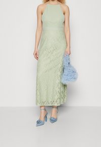 Ljusgrön spetsklänning med halterneck, lagerdesign. Kombinerad med en ljusblå volangväska och blå högklackade skor med blommor som dekoration.